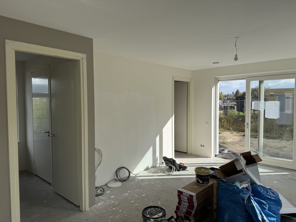 Kale woonkamer van een nieuw opgeleverd huis met schuifpui, binnendeuren en emmers verf op de betonvloer