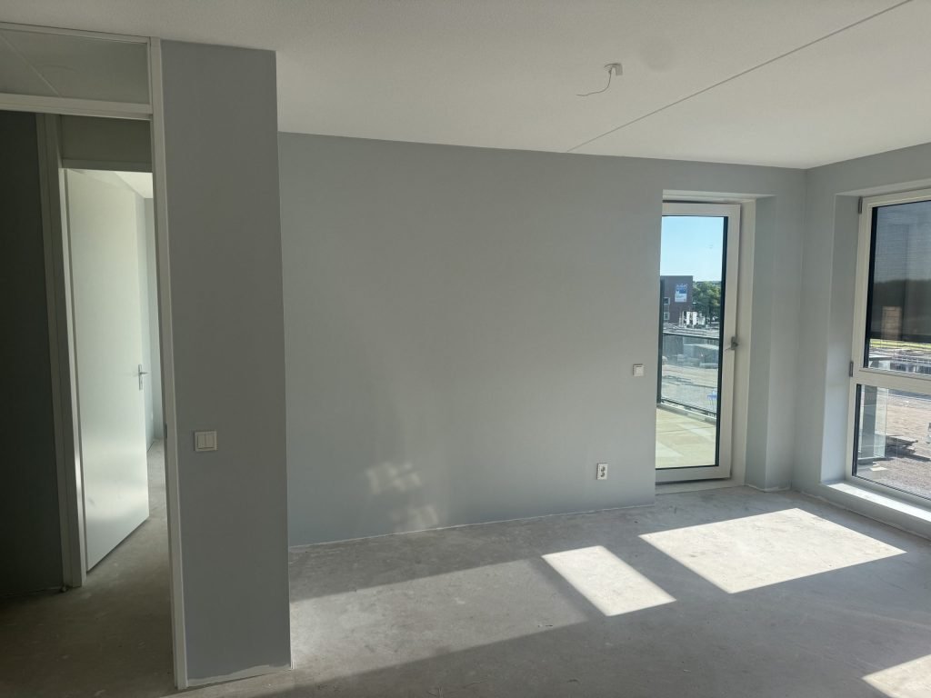 Appartement in aanbouw met lichtgrijs gesausde wanden, een balkondeur en zonlicht op de betonvloer