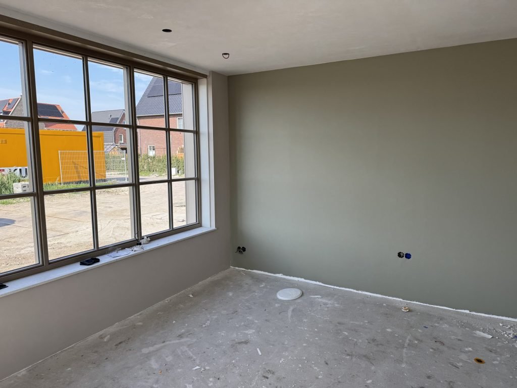 Nieuwbouwkamer met groot sprossenraam en een olijfgroen gesausde wand, buiten nog een bouwkeet