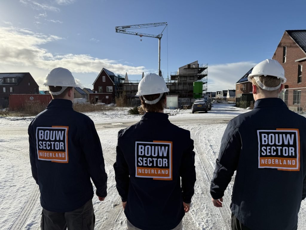 Drie vakmensen van Bouwsector Nederland met witte helm op de rug gezien, voor een nieuwbouwproject met bouwkraan in de sneeuw
